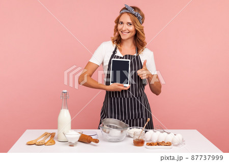 Portrait of attractive young adult female in apron cooking bakery, holding blank tablet in hand and showing thumb up, looking at camera with smile. Indoor studio shot isolated on pink background. 87737999