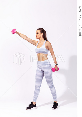 Full length side view athletic woman in white sports top and tights raising hands with dumbbells, pumping up muscles and developing strength. Indoor studio shot isolated on gray background Full length side view athletic woman in white sports top and tights raising hands with dumbbells, pumping up muscles and developing strength. Indoor studio shot isolated on gray background 87738241