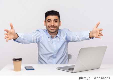 Come into my arms. Excited happy friendly businessman sitting office workplace, stretching hands to camera, going to embrace, welcoming on reception. indoor studio shot isolated on white background Come into my arms. Excited happy friendly businessman sitting office workplace, stretching hands to camera, going to embrace, welcoming on reception. indoor studio shot isolated on white background 87738274