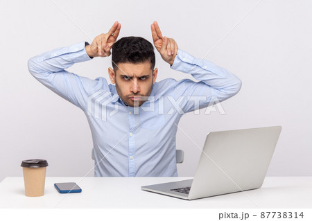 Conflict at work. Aggressive man employee sitting office workplace and showing bull horn gesture, threatening to attack, looking angrily at camera. indoor studio shot isolated on white background 87738314