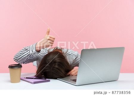 I am fine. Extremely tired sleepy woman lying on table with laptop and cup of coffee showing thumbs up, exhausted with overwork, vitamin deficiency. Indoor studio shot isolated on pink background 87738356