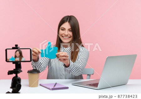 Excited female blogger showing blue thumbs up template on smartphone camera, keeping online communication with subscribers, asking for rate. Indoor studio shot isolated on pink background Excited female blogger showing blue thumbs up template on smartphone camera, keeping online communication with subscribers, asking for rate. Indoor studio shot isolated on pink background 87738423