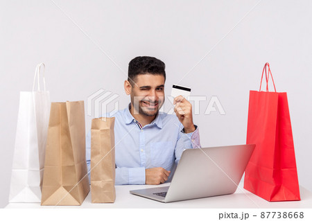 E-commerce, internet shopping. Cheerful man sitting at desk with laptop, surrounded by packages, showing bank card in his hand and winking, smiling playful. studio shot isolated on white background 87738668