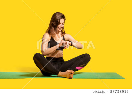 Cheerful sporty girl sitting on gym mat, looking time, touching smartwatch display to check pulse in fitness tracker, monitoring heart beat, wearable app. Healthy lifestyle, isolated studio shot Cheerful sporty girl sitting on gym mat, looking time, touching smartwatch display to check pulse in fitness tracker, monitoring heart beat, wearable app. Healthy lifestyle, isolated studio shot 87738915