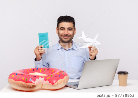 Cheerful man holding paper airplane and passport in hands, sitting in office workplace with rubber ring on desk, planning travel vacation, weekend trip. studio shot isolated on white background Cheerful man holding paper airplane and passport in hands, sitting in office workplace with rubber ring on desk, planning travel vacation, weekend trip. studio shot isolated on white background 87738952