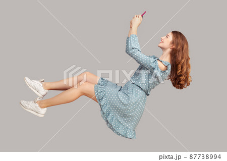 Hovering in air. Smiling girl ruffle dress levitating with mobile phone, reading message chatting happy joyful in social network online, surfing web while flying. indoor studio shot isolated on gray 87738994