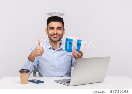 Congratulations. Joyful angelic businessman with nimbus on head showing gift box and thumbs up, sitting at laptop workplace, greeting with professional holidays. studio shot isolated, white background 87739148