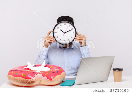 Time to rest. Unknown man hiding face behind clock, sitting in office workplace with rubber ring passport and paper airplane on desk, employee asking for break, relax and travel vacation. studio shot 87739199