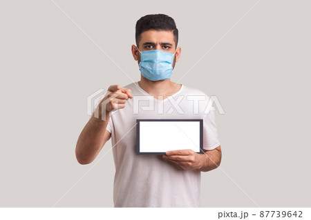 StayAtHome. Man in protective mask holding Stay at home inscription and pointing to camera, warning of coronavirus quarantine, Covid-19 epidemic, preventive measures for spread of infectious disease. 87739642