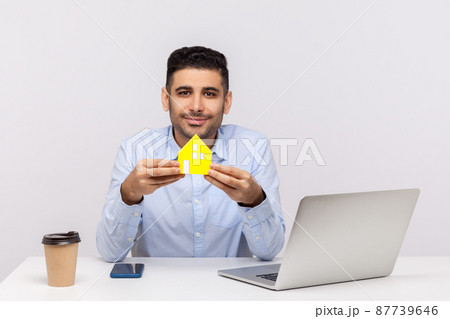 Portrait of real estate agent man sitting in office workplace and holding paper house, smiling friendly, advertisement of purchase or rental housing. indoor studio shot isolated on white background Portrait of real estate agent man sitting in office workplace and holding paper house, smiling friendly, advertisement of purchase or rental housing. indoor studio shot isolated on white background 87739646
