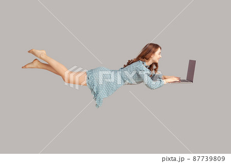 Hovering in air. Happy cheerful girl ruffle dress levitating with laptop, typing keyboard, reading good news message on computer while flying in mid-air. studio shot isolated on gray background 87739809