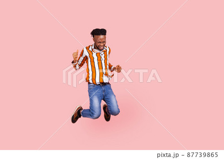 Full length happy positive african man with dreadlocks in stylish bright shirt highly jumping on trampoline, having fun, enjoying like in childhood. Indoor studio shot isolated on pink background Full length happy positive african man with dreadlocks in stylish bright shirt highly jumping on trampoline, having fun, enjoying like in childhood. Indoor studio shot isolated on pink background 87739865