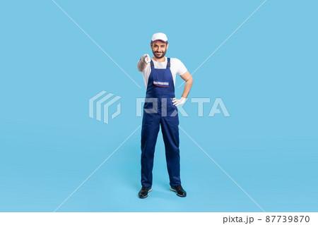 Full length happy courier in overalls, cap and protective gloves standing, pointing finger to camera, choosing you. Profession of service industry, loader or post delivery. indoor studio shot isolated Full length happy courier in overalls, cap and protective gloves standing, pointing finger to camera, choosing you. Profession of service industry, loader or post delivery. indoor studio shot isolated 87739870