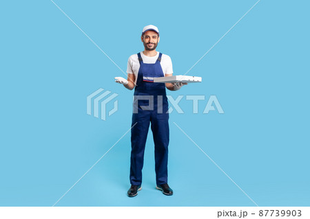 Full length happy courier in workwear and protective gloves carrying pizza box, holding empty space on palm for advertise image. Fast food delivery service. studio shot isolated on blue background Full length happy courier in workwear and protective gloves carrying pizza box, holding empty space on palm for advertise image. Fast food delivery service. studio shot isolated on blue background 87739903