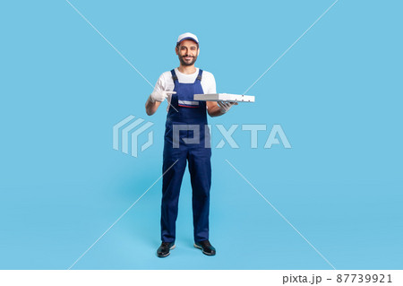 Full length positive professional courier in workwear and protective gloves pointing at pizza box, smiling friendly to camera. Fast food delivery service. studio shot isolated on blue background 87739921