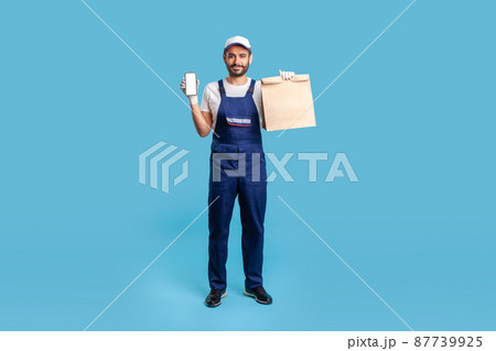 Full body portrait of expert courier in blue uniform holding groceries bag and mobile phone with mock up blank display for professional delivery advertise, online food purchase. studio shot isolated 87739925