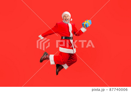 Extremely happy santa claus holding gift box jumping high and looking at camera with toothy smile, satisfied with winter holidays. Indoor studio shot isolated on red background Extremely happy santa claus holding gift box jumping high and looking at camera with toothy smile, satisfied with winter holidays. Indoor studio shot isolated on red background 87740006