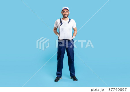 Full length handyman, building worker in uniform holding paper house. Mover guy offering help in relocation, home repair services and maintenance. indoor studio shot isolated on blue background 87740097