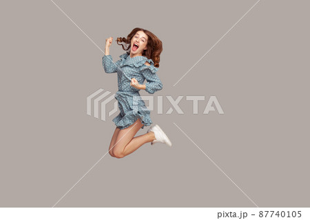 Delighted enthusiastic girl ruffle dress flying mid-air with raised fists shouting for joy, jumping trampoline looking at camera extremely happy, celebrating success. studio shot isolated on gray 87740105