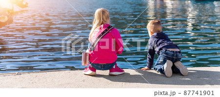 Back view two cute little curious sibling kids sitting on concrete embankment pier near sea lake bank and watching fish ship yacht boat marina. Children enjoy have fun playing outdoors at summer day 87741901