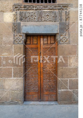 Wooden ornate door with geometrical engraved patterns on external old decorated bricks stone wall Wooden ornate door with geometrical engraved patterns on external old decorated bricks stone wall 87744138