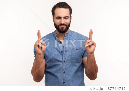Hopeful dreamy man with beard in blue shirt crossing fingers and closing eyes making wish, believing in miracle, hope in success. Indoor studio shot isolated on white background Hopeful dreamy man with beard in blue shirt crossing fingers and closing eyes making wish, believing in miracle, hope in success. Indoor studio shot isolated on white background 87744712