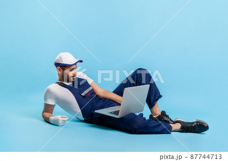 Full length handsome expert handyman in uniform lying sexy posing on camera, reading message on laptop. Profession of online customer support, contact center of repair service. studio shot isolated Full length handsome expert handyman in uniform lying sexy posing on camera, reading message on laptop. Profession of online customer support, contact center of repair service. studio shot isolated 87744713