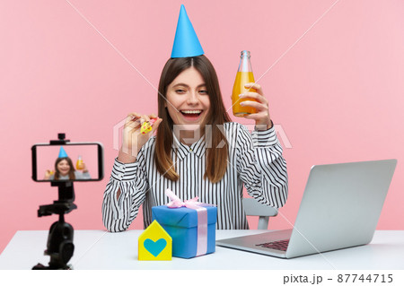 Cheerful optimistic woman blogger celebrating birthday with subscribers, posing at smartphone camera, recording video, online streaming. Indoor studio shot isolated on pink background Cheerful optimistic woman blogger celebrating birthday with subscribers, posing at smartphone camera, recording video, online streaming. Indoor studio shot isolated on pink background 87744715