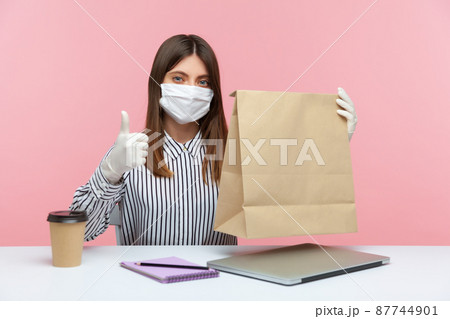 Satisfied woman office employee sitting at workplace, feeling safe healthy with protective face mask and gloves, holding paper bag, showing thumbs up. Excellent food delivery in covid-19 quarantine Satisfied woman office employee sitting at workplace, feeling safe healthy with protective face mask and gloves, holding paper bag, showing thumbs up. Excellent food delivery in covid-19 quarantine 87744901