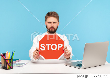 Serious bearded man office worker holding red stop sign board sitting at workplace with laptop, rejecting sexual harassment at work. Indoor studio shot isolated on blue background 87744933
