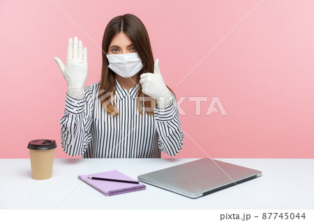 Good protection against coronavirus infection. Woman office employee sitting at workplace, feeling safe healthy with protective mask and gloves during covid-19 quarantine, showing thumbs up. indoor Good protection against coronavirus infection. Woman office employee sitting at workplace, feeling safe healthy with protective mask and gloves during covid-19 quarantine, showing thumbs up. indoor 87745044