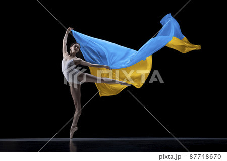 Young graceful classic ballerina dancing with cloth painted in blue and yellow colors of Ukraine flag on dark studio background. Art, peace, freedom and rights 87748670