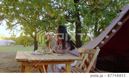 A woman sits at a wooden table in nature drinking tea. Outdoors A woman sits at a wooden table in nature drinking tea. Outdoors 87748855