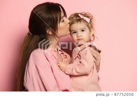 Kiss. Portrait of young woman and little girl, mother and daughter isolated on pink studio background. Mother's Day celebration. Concept of family, childhood, motherhood Kiss. Portrait of young woman and little girl, mother and daughter isolated on pink studio background. Mother's Day celebration. Concept of family, childhood, motherhood 87752050