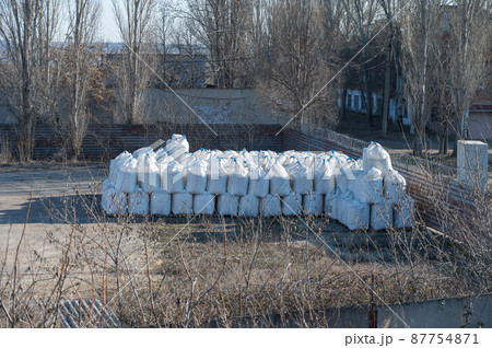 A stack of large bags outdoors. Industrial warehouse. Trees without leaves around the perimeter of the warehouse. A stack of large bags outdoors. Industrial warehouse. Trees without leaves around the perimeter of the warehouse. 87754871