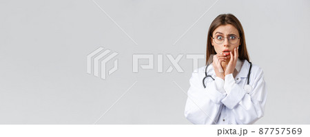 Healthcare workers, medicine, insurance and covid-19 pandemic concept. Shocked scared and concerned female doctor in white scrubs and glasses listening to scary news, gasping, hold hands near lips 87757569