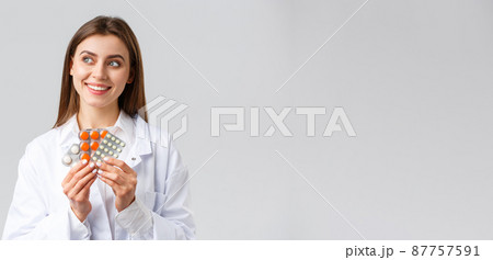 Pharmacy, healthcare workers, insurance and hospitals concept. Thoughtful smiling and pleased female doctor in white scrubs, look away thinking, holding medicine, pills or vitamins Pharmacy, healthcare workers, insurance and hospitals concept. Thoughtful smiling and pleased female doctor in white scrubs, look away thinking, holding medicine, pills or vitamins 87757591