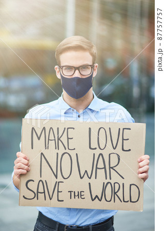 Vertical of young male activist wearing black protective mask holding banner or poster and looking at camera while protesting against the war outdoors, standing on the road 87757757
