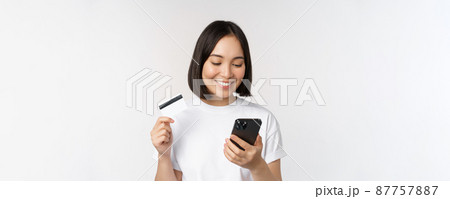 Online shopping. Happy asian woman using credit card and smartphone app, paying on website via mobile phone, white background Online shopping. Happy asian woman using credit card and smartphone app, paying on website via mobile phone, white background 87757887