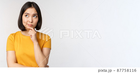 Image of thinking asian woman, looking aside and pondering, making decision, standing in yellow tshirt over white background 87758116