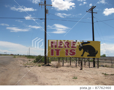 Along a desert road, a yellow billboard sign reads Here It Is with a rabbit's silhouette 87762460
