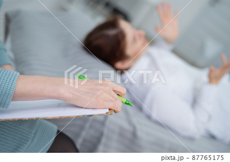 A caucasian woman lies on a couch and expresses her feelings, while a psychologist makes notes on a tablet.  87765175