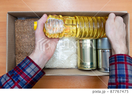 Man in a shirt stacks food in case of emergency, natural disasters, self-isolation, pandemics, covid 19. Food with a long shelf life. Canned food, oil, rice, buckwheat. Help for the starving Man in a shirt stacks food in case of emergency, natural disasters, self-isolation, pandemics, covid 19. Food with a long shelf life. Canned food, oil, rice, buckwheat. Help for the starving 87767475
