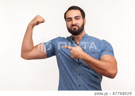 Self confident egotistical bearded man in blue shirt pointing finger at his arm biceps, proud and satisfied with muscular build, leadership skills. Indoor studio shot isolated on white background 87768029