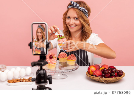 Food vlogger holding small cake decorating with cream and cherry, looking at camera on smart phone on tripod and smiling, recording video for vlog. Indoor studio shot isolated on pink background. 87768096