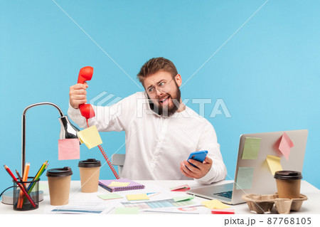 Busy overworked man sitting at workplace all covered with sticky notes and holding three phones in hands, answering calls, service center. Indoor studio shot isolated on blue background. Busy overworked man sitting at workplace all covered with sticky notes and holding three phones in hands, answering calls, service center. Indoor studio shot isolated on blue background. 87768105