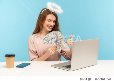 Recommending big money profit. Lovely angelic rich woman with nimbus holding dollar banknotes and showing thumbs up while talking video call on laptop. indoor studio shot isolated on blue background 87768158