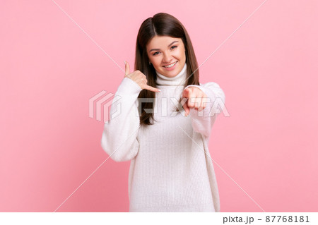 Smiling positive young adult female showing call me gesture with hand and pointing finger at camera, wearing white casual style sweater. Indoor studio shot isolated on pink background. Smiling positive young adult female showing call me gesture with hand and pointing finger at camera, wearing white casual style sweater. Indoor studio shot isolated on pink background. 87768181