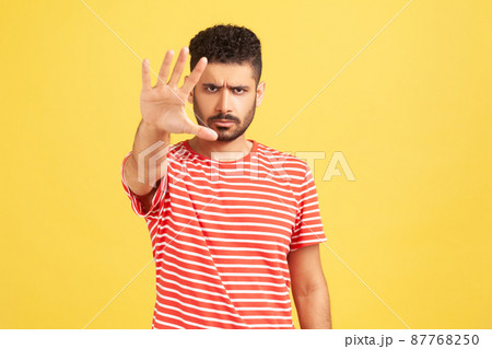 Serious concerned man with beard in striped t-shirt making stop gesture showing palm of hand, conflict prohibition warning about danger, stop bullying. Indoor studio shot isolated on yellow background 87768250
