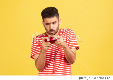 Addicted nervous bearded man in red striped t-shirt grimacing holding joystick, playing video games online, virtual competition. Indoor studio shot isolated on yellow background 87768332